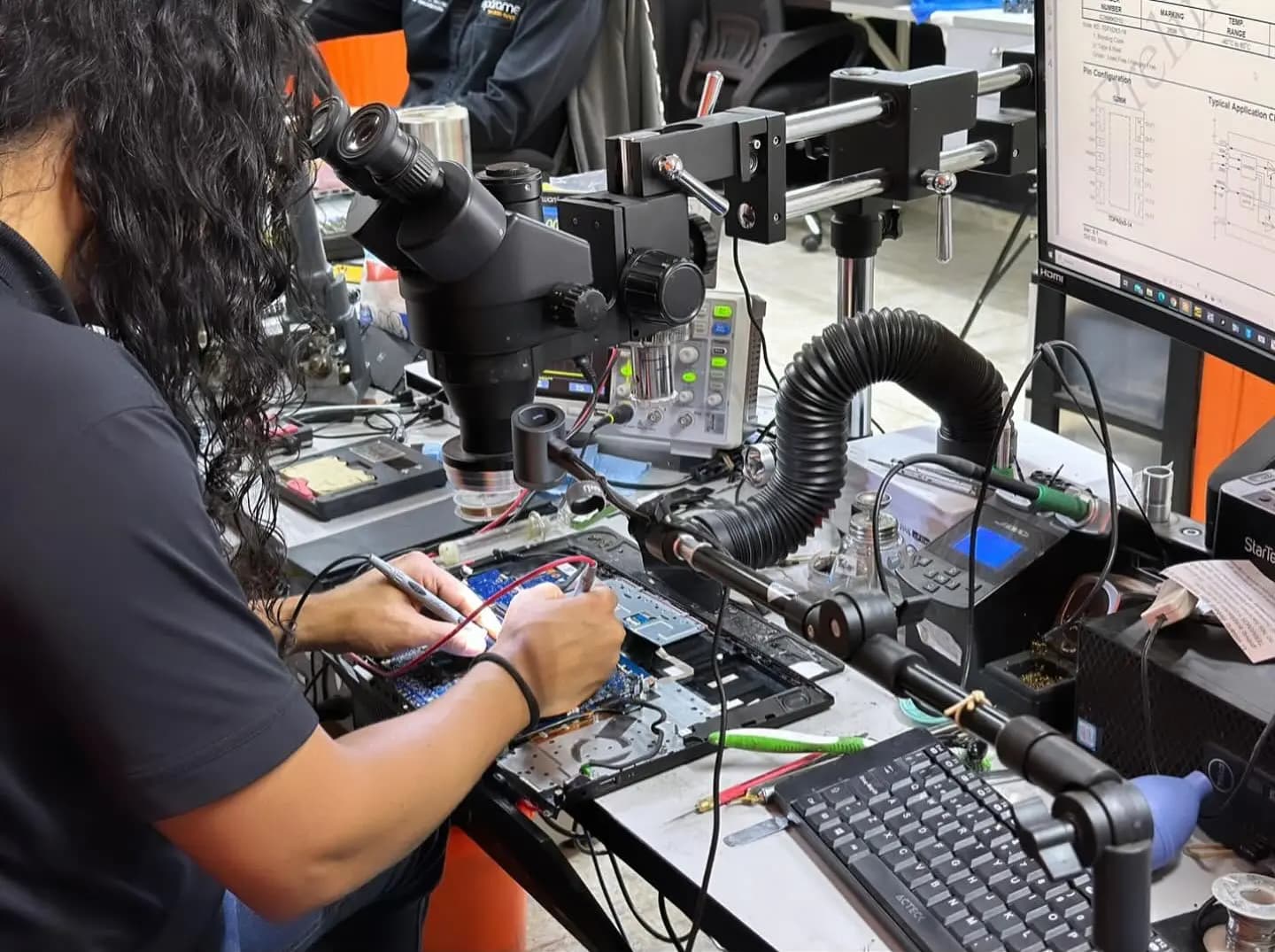 Técnica de Repárame midiendo la tarjeta madre de una laptop bajo el microscopio, con esquema eléctrico en pantalla